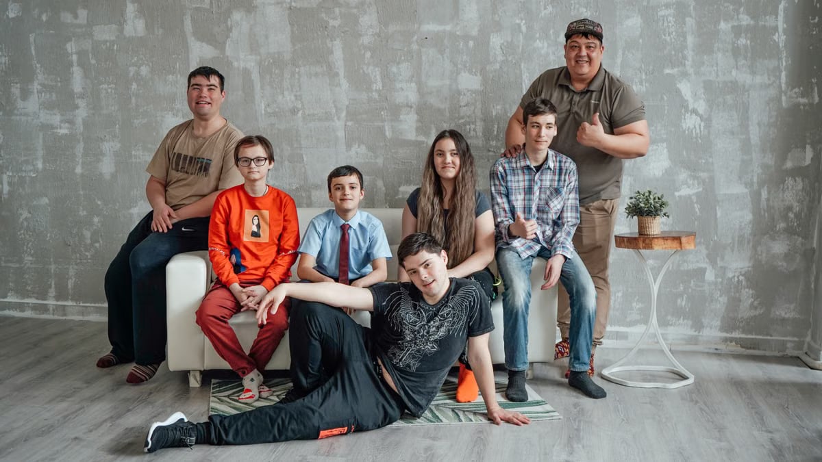 Group of seven people posing casually in a stylish indoor setting with a concrete wall background.